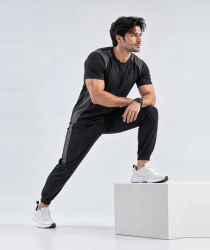 A man in a black training tee and joggers with a contrast side panel, stretching with one foot raised on a block.