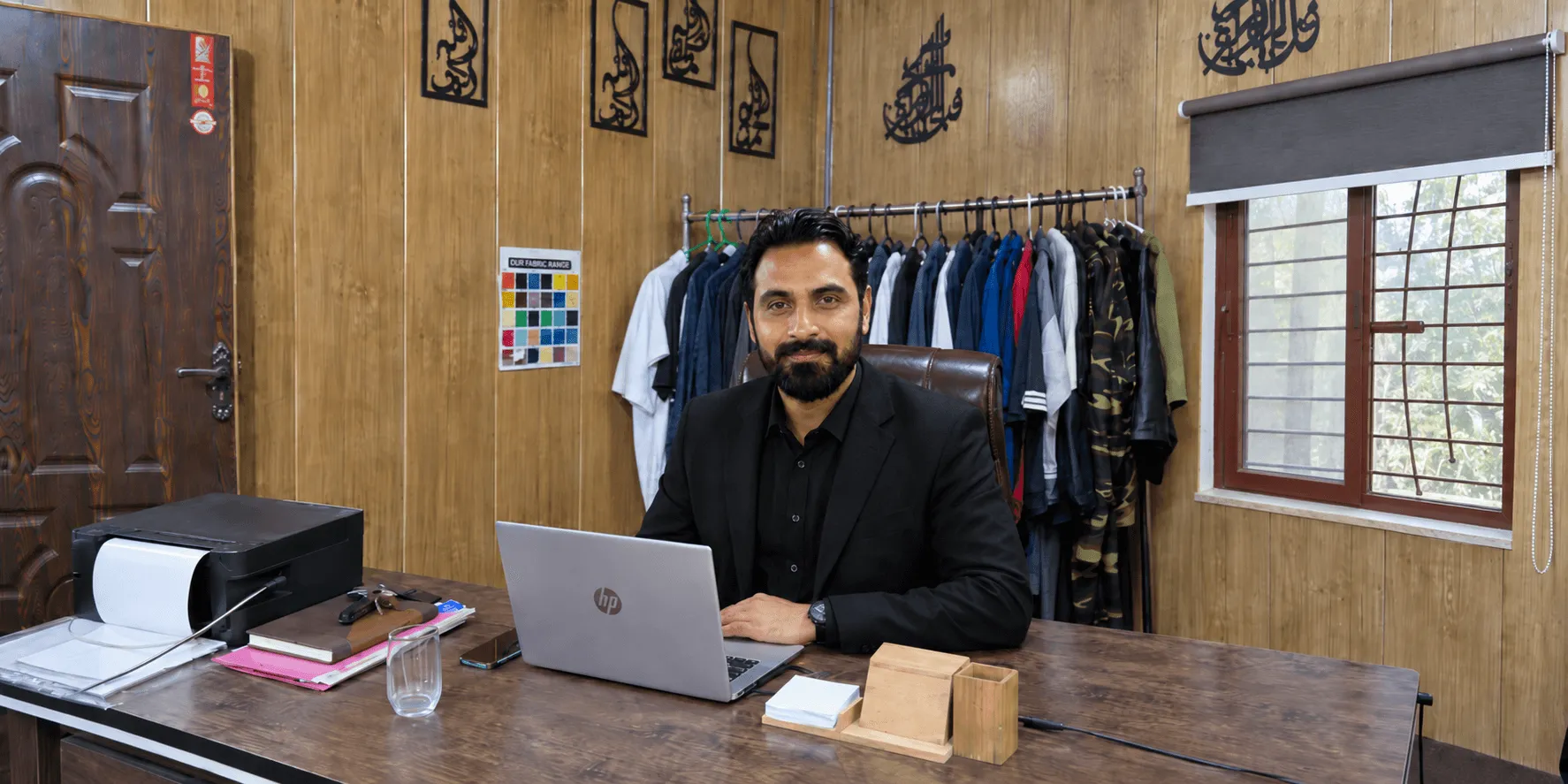 Khaleeq Ur Rehman at his desk in the Sialkot office, with the fabric colour card and a rail of sample garments on the wall behind him.