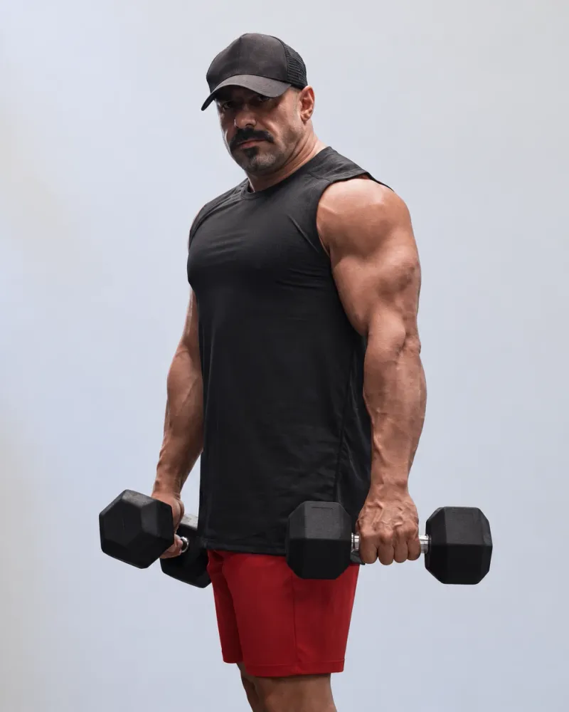 A model in a black sleeveless training top with a dropped armhole and red athletic shorts, holding a dumbbell in each hand, against a pale studio backdrop.