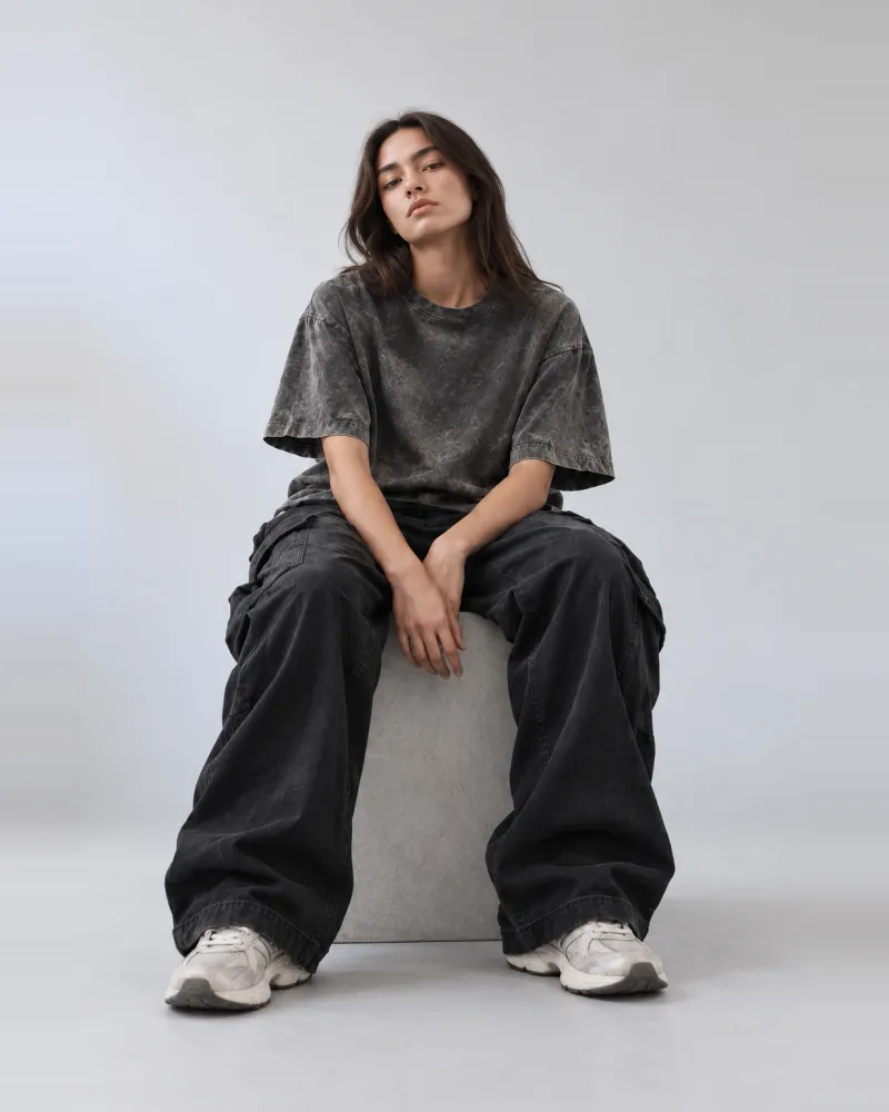 A seated model in oversized wide-leg washed-black cargo trousers with deep side pockets and a heavy drape, worn with an acid-wash grey t-shirt and white trainers.