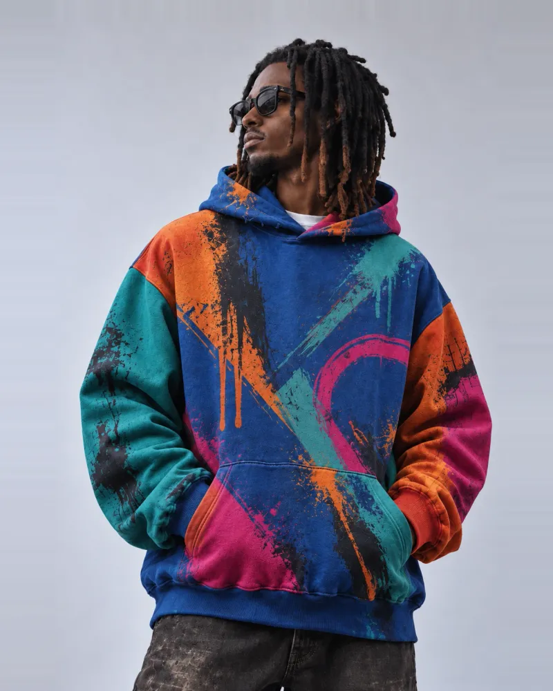 A model in an oversized cobalt-blue hoodie printed edge to edge with a paint-splatter graphic in orange, teal and magenta, with colour-blocked sleeves and a kangaroo pocket.