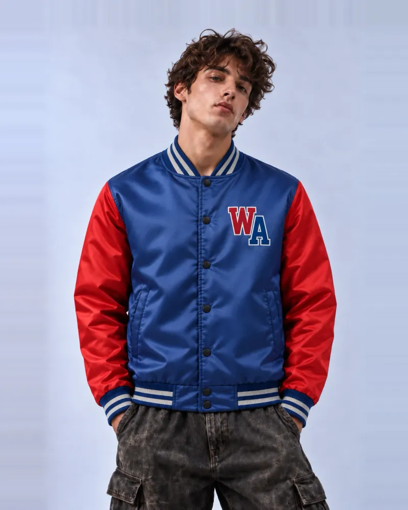 A model in a royal blue varsity jacket with red raglan sleeves, striped ribbed collar, cuffs and hem, a snap front, and a red and blue "WA" letter patch on the chest, worn with washed grey cargo trousers.