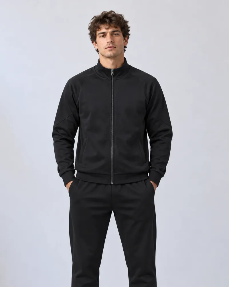 A model in a plain black tracksuit — a zip-through track jacket with a stand collar and raglan sleeves, worn with matching tapered joggers — against a pale studio backdrop.