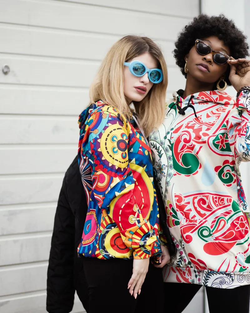 Two models in bold, full-coverage all-over printed pullover hoodies with contrasting patterns.